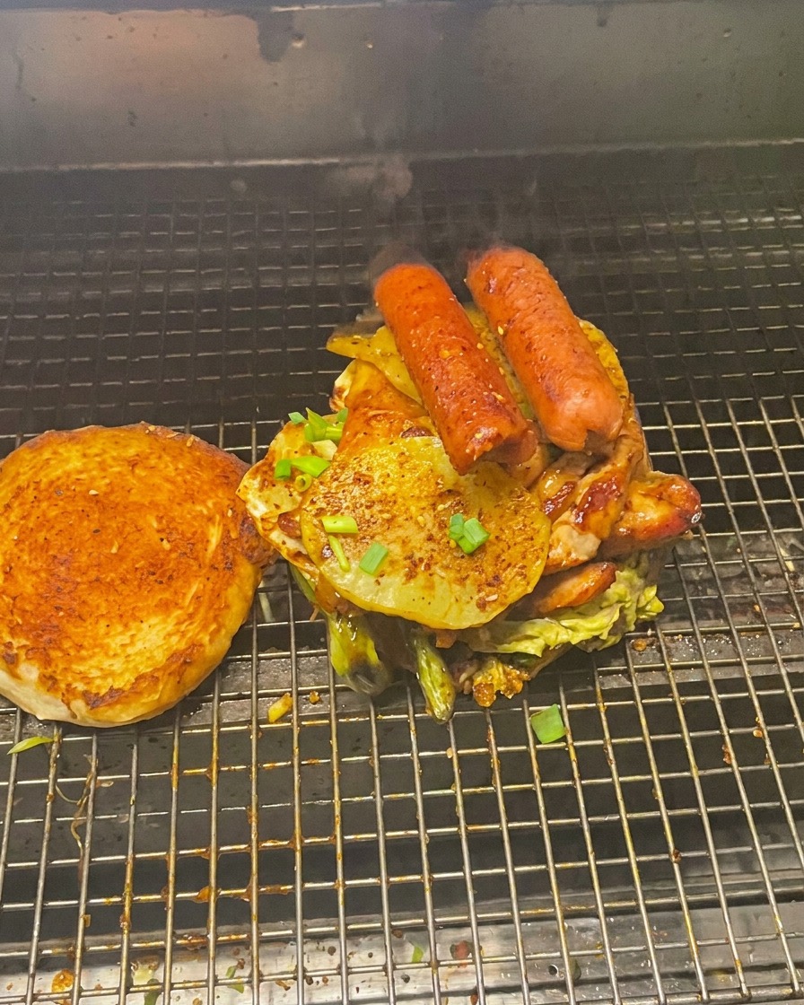 Une tour imposante de viandes et légumes grillés sur un pain, avec le chapeau toasté posé à côté sur le grill.