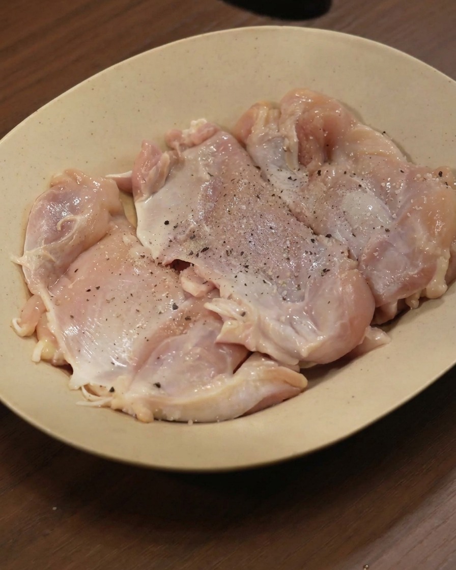 Hauts de cuisses de poulet crus sur une assiette, saupoudrés de poivre noir et dépices moulues.