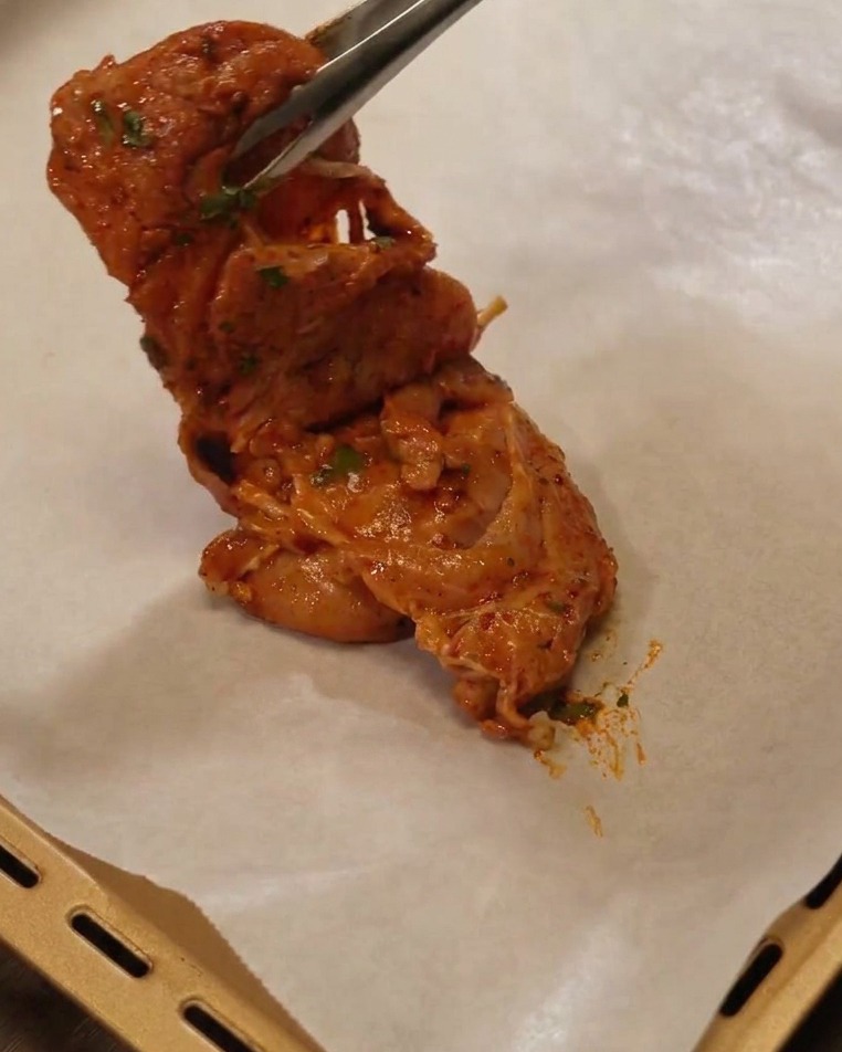 Une pince métallique déposant un haut de cuisse mariné sur une plaque de cuisson tapissée de papier sulfurisé.