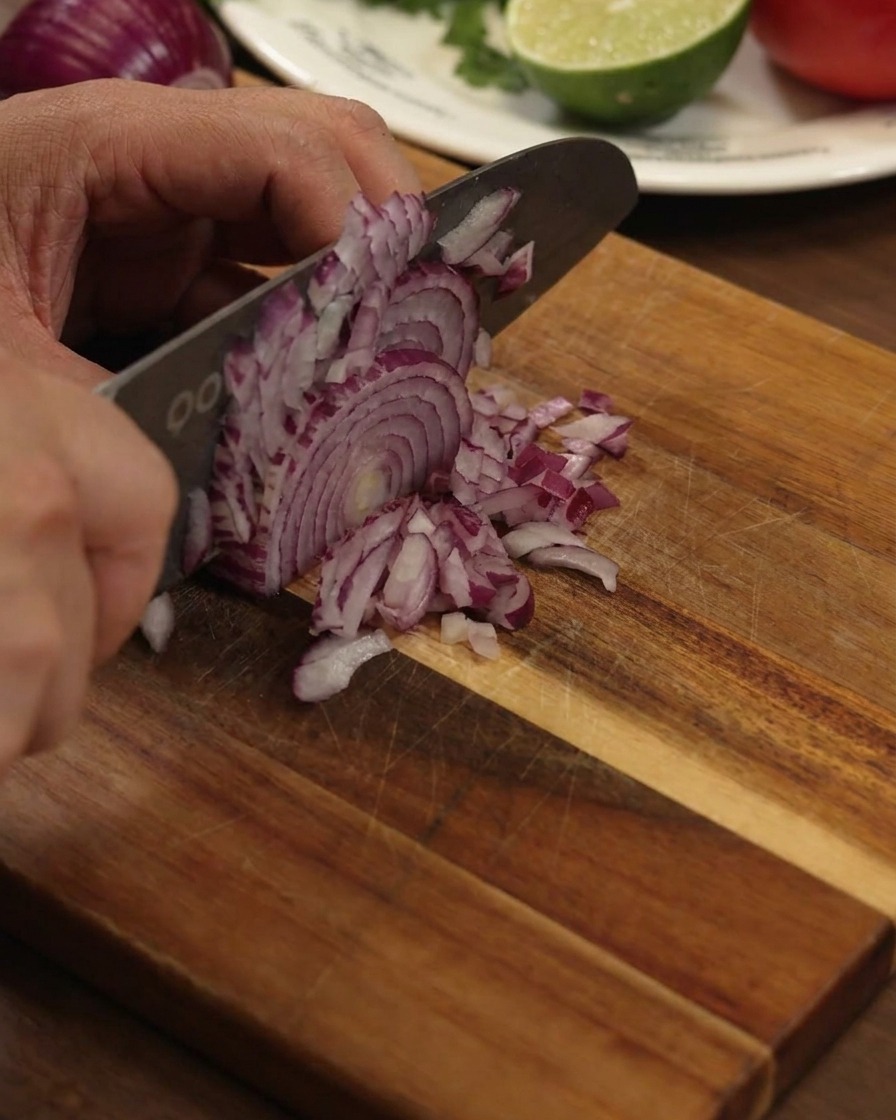 Une main utilisant un grand couteau de cuisine pour émincer finement un oignon rouge sur une planche en bois.
