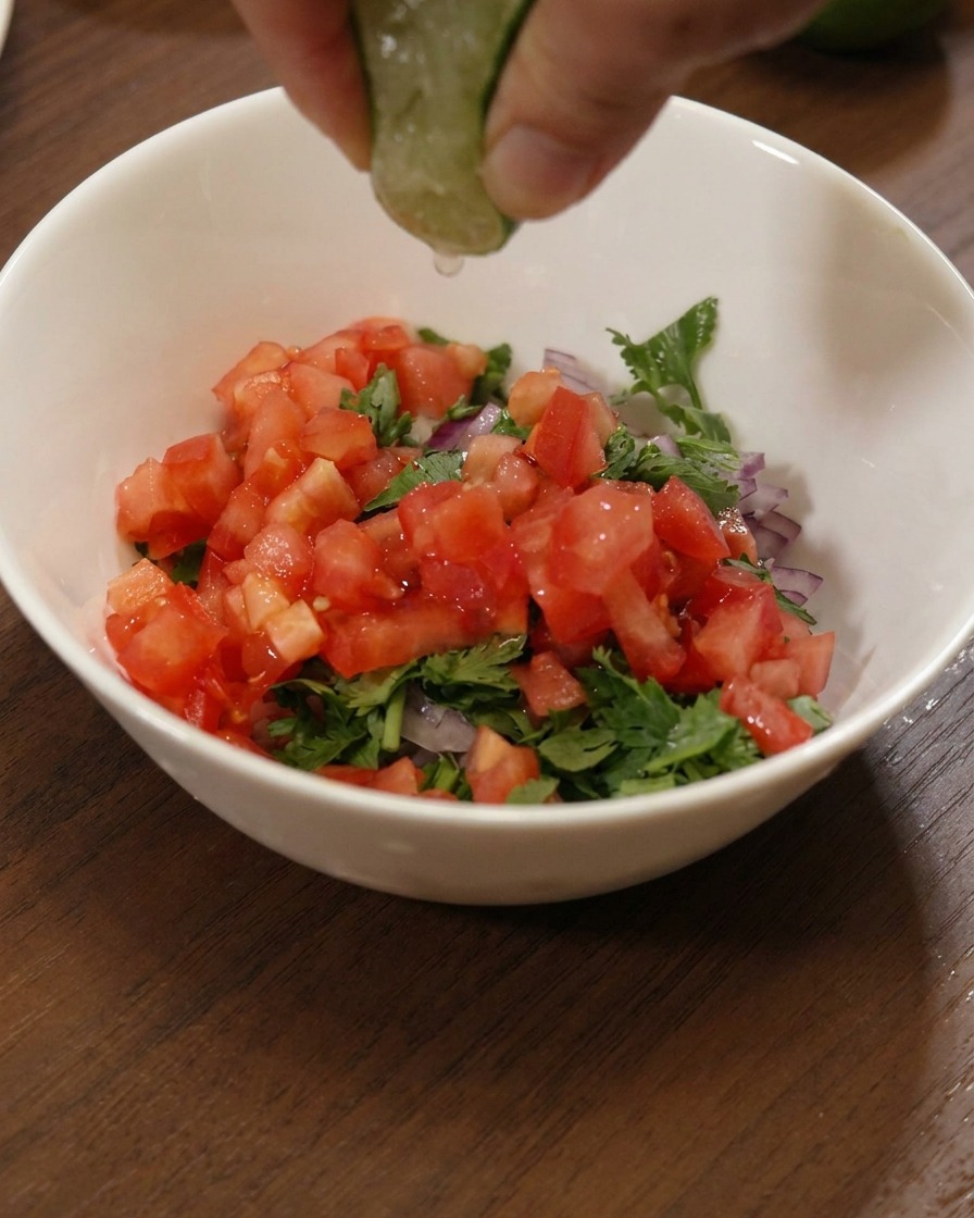Une main pressant un quartier de citron vert frais sur un bol de tomates, doignons et de coriandre.