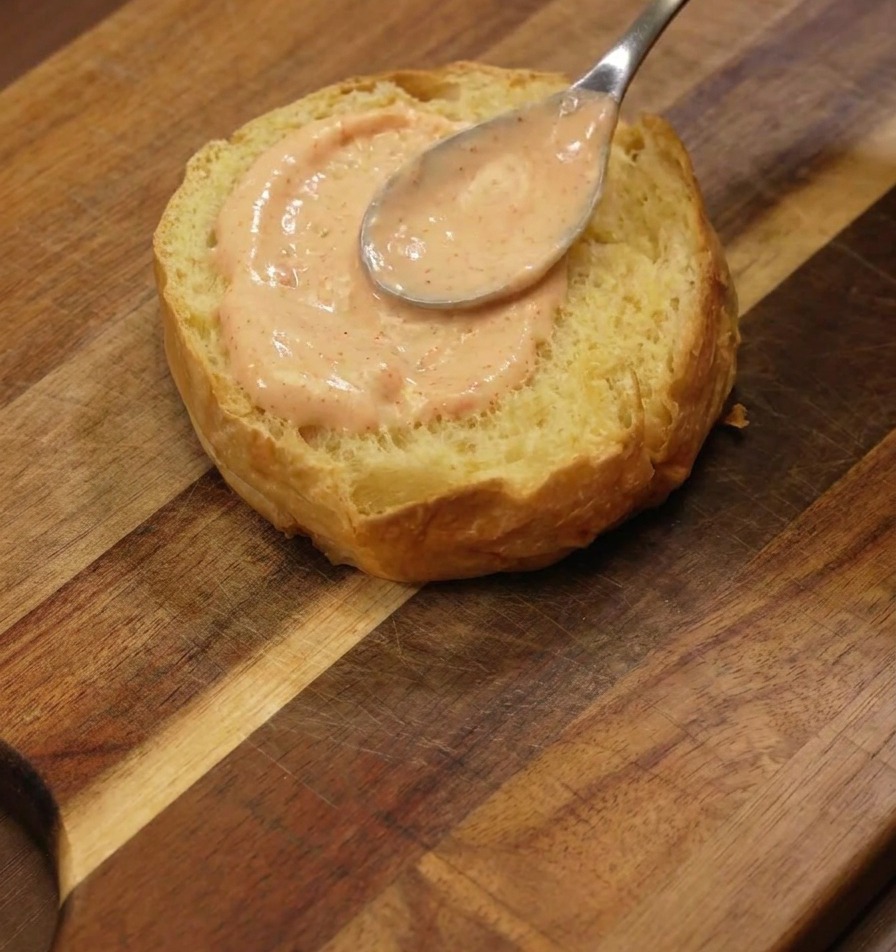Une cuillère en argent étalant une couche épaisse de sauce orange sur la face toastée dun pain brioché sur une planche en bois.