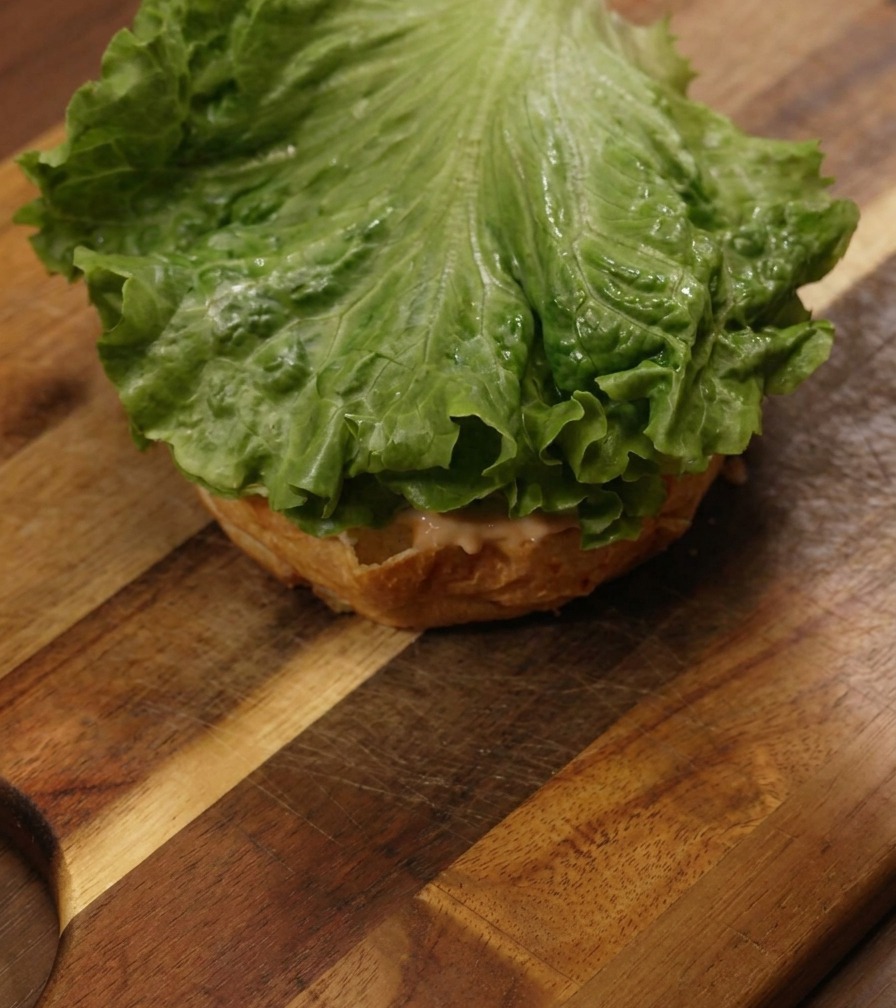 Une feuille de laitue verte croquante posée sur un pain burger nappé de sauce sur une planche en bois.
