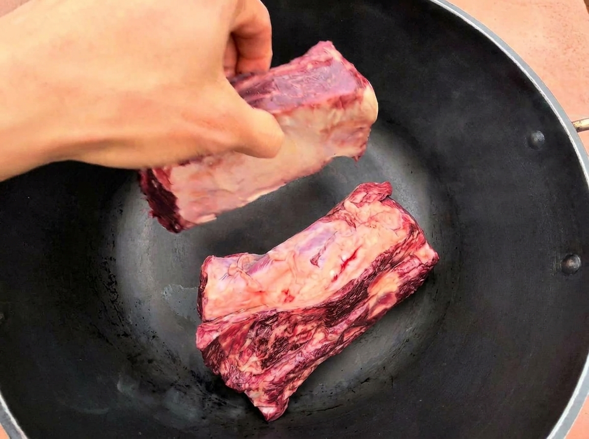 Une main déposant deux grandes côtes de bœuf crues dans une poêle noire bien chaude.