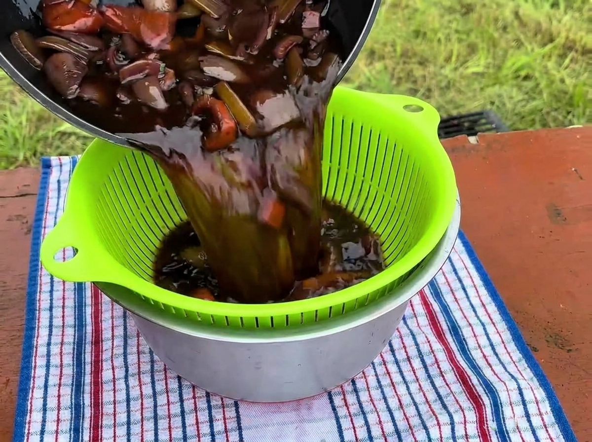 Le jus de cuisson foncé et les légumes versés dans une passoire verte au-dessus dun bol en inox.