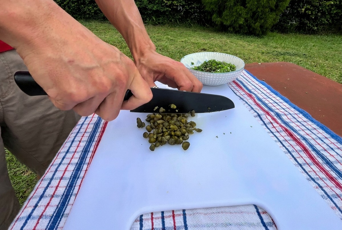 Gros plan sur un couteau de chef hachant finement des câpres vertes sur une planche à découper.