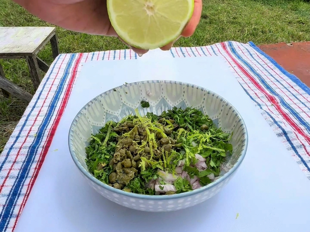Une main pressant un demi-citron vert au-dessus dun bol en céramique rempli dherbes hachées, de câpres, déchalotes et de zestes.