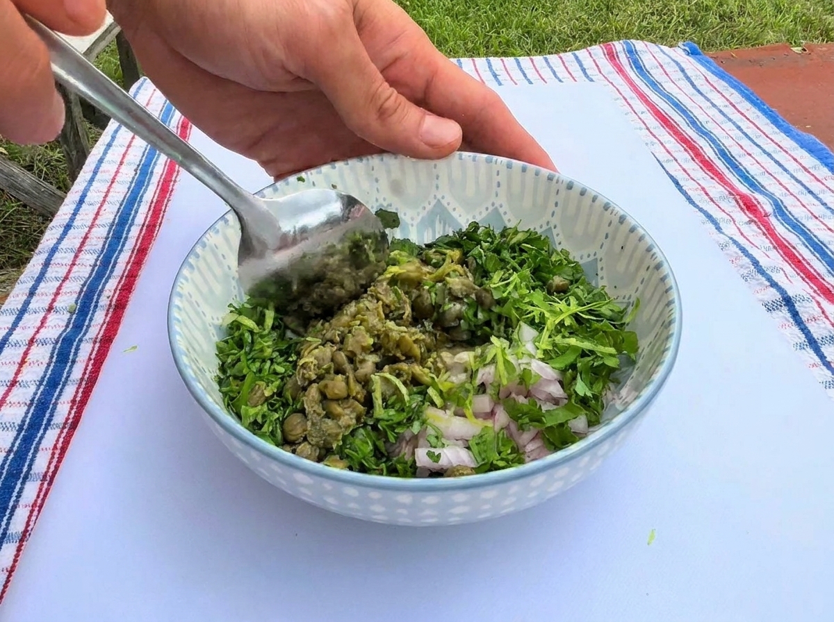Mélange dherbes vertes hachées, de câpres et déchalotes avec une cuillère dans un bol en céramique décoré.