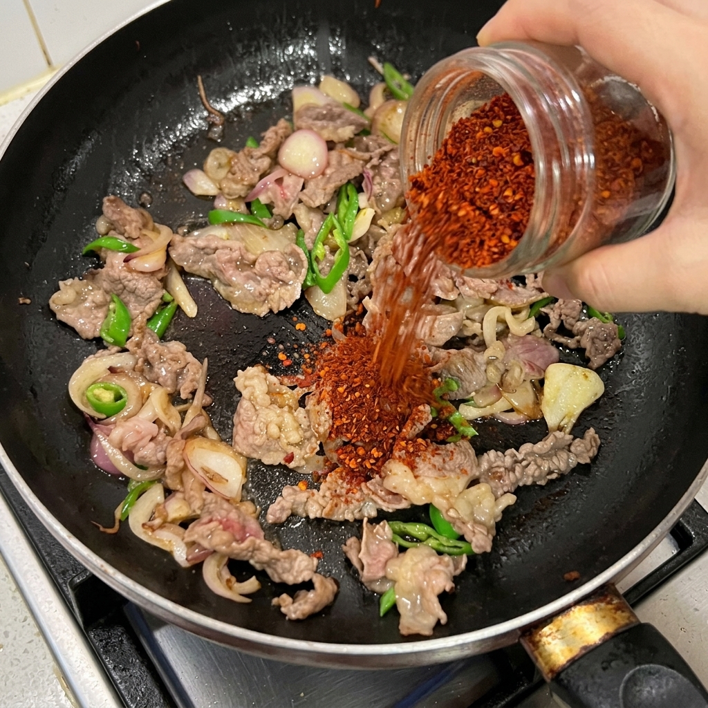 Du piment rouge en poudre versé depuis un pot en verre sur le bœuf émincé, les oignons et les piments verts sautant dans une poêle noire.