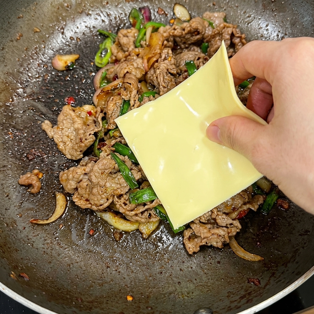 Une main déposant une tranche carrée de fromage jaune sur un mélange chaud de bœuf sauté et de piments verts dans une poêle.