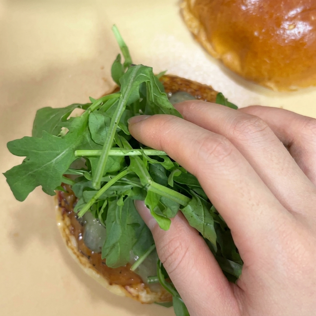 Une main déposant un bouquet de feuilles de roquette fraîche sur des cornichons dans un pain à burger.