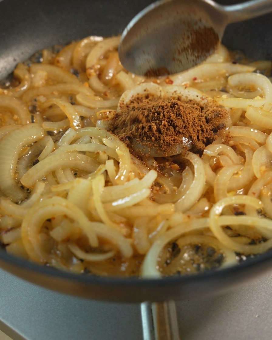 Tranches doignons sautées dans une poêle avec une cuillère ajoutant de la cassonade et des condiments liquides.