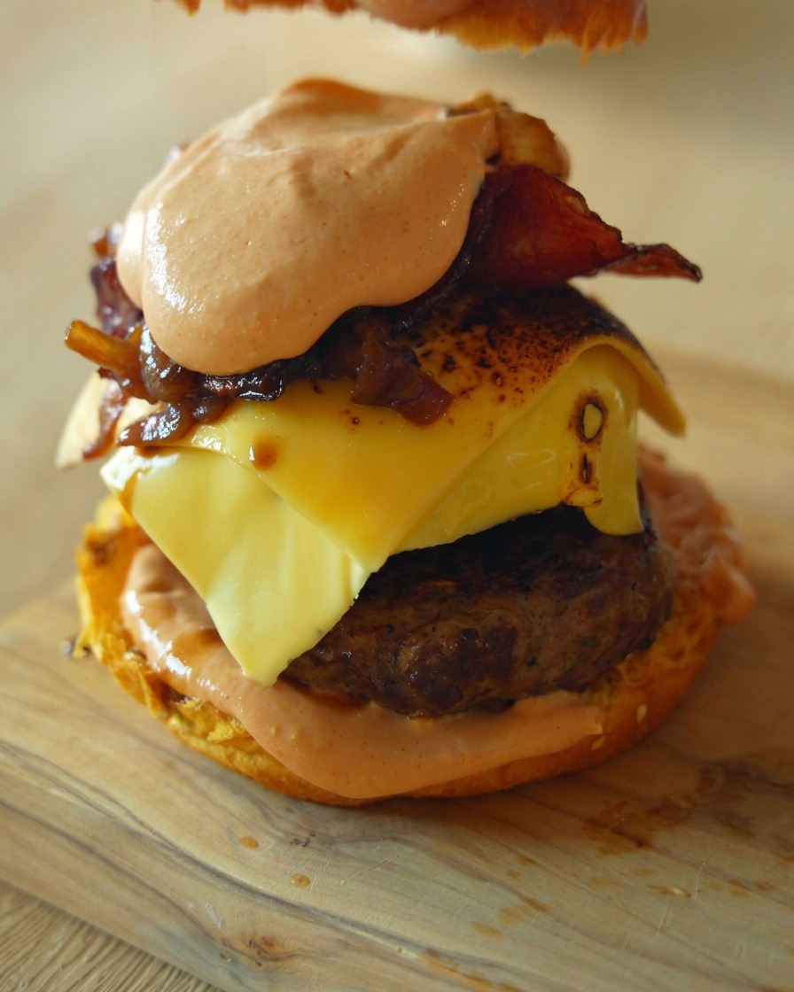 Main posant le chapeau du pain sur un burger gourmet imposant avec steak, fromage, oignons et sauce.