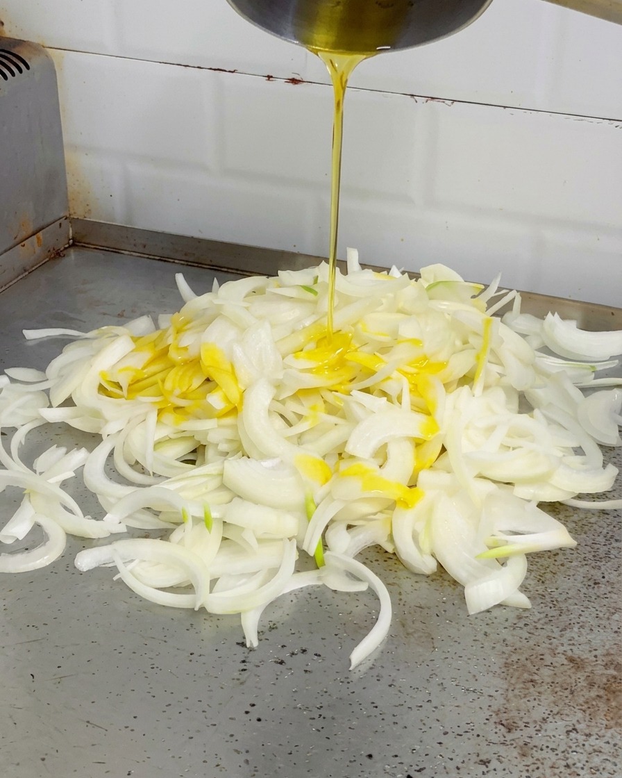 Du beurre jaune fondu versé depuis une petite casserole sur un tas doignons blancs sur une plancha.