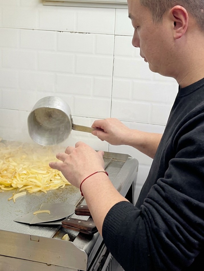 Un chef versant de leau avec une petite louche en métal sur des oignons qui ramollissent sur une plaque de cuisson professionnelle.