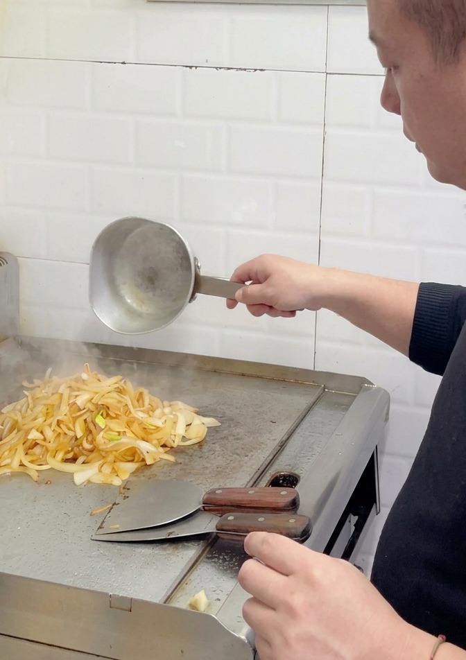 Un chef ajoutant plus deau à un tas doignons légèrement dorés et partiellement caramélisés sur une plaque.