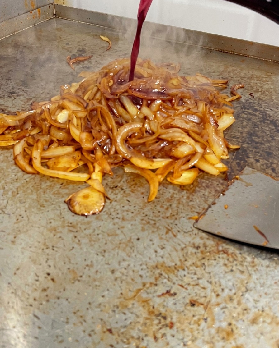 Du vin rouge versé dune bouteille sur des oignons dun brun profond sur une plancha.