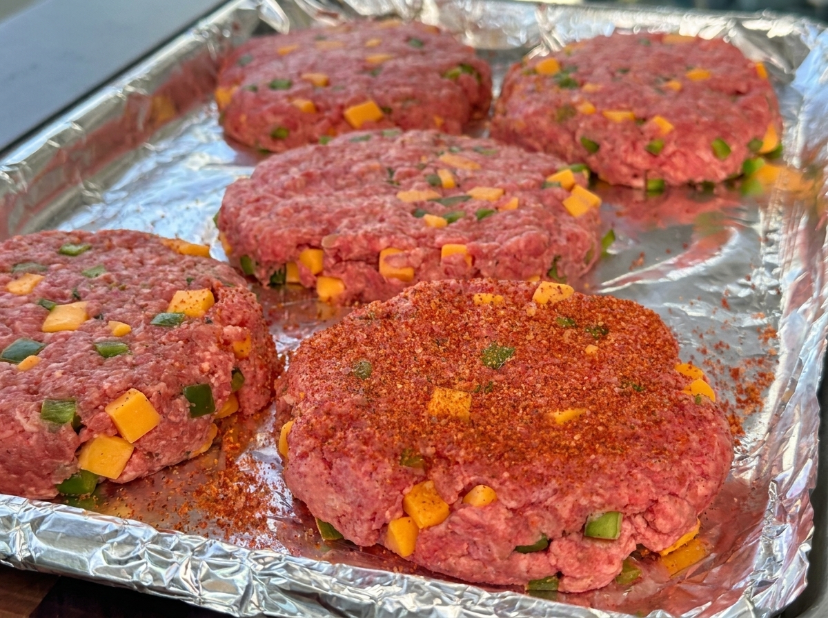 Steaks de bœuf crus façonnés laissant apparaître les dés de fromage et de poivron, saupoudrés dépices BBQ sur une plaque.
