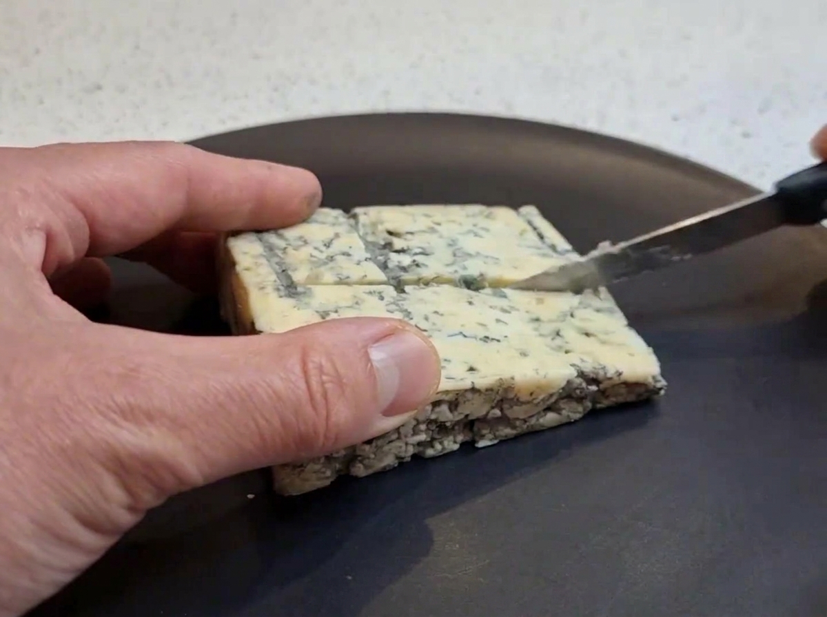 Une main coupant un morceau carré de fromage bleu en petits dés sur une assiette sombre à laide dun petit couteau.