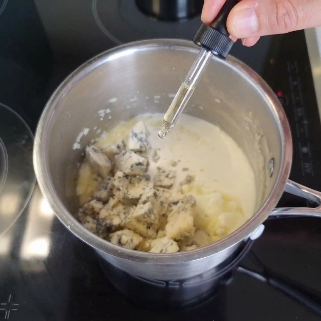 Une main utilisant une pipette en verre pour verser de la sauce Worcestershire sombre dans une casserole contenant de la crème et des dés de bleu.