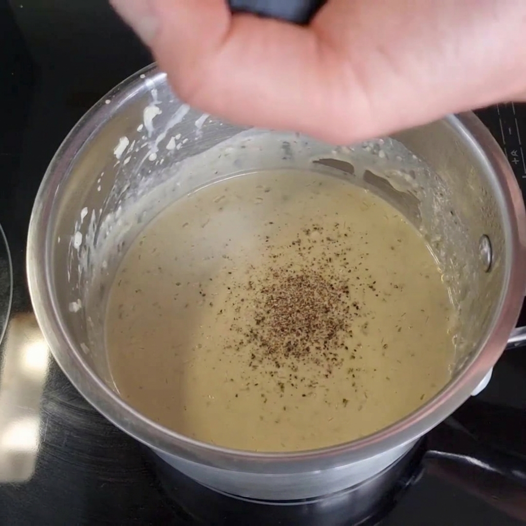 Une main actionnant un moulin à poivre pour moudre du poivre noir frais directement dans une sauce crémeuse à lintérieur dune casserole en inox.