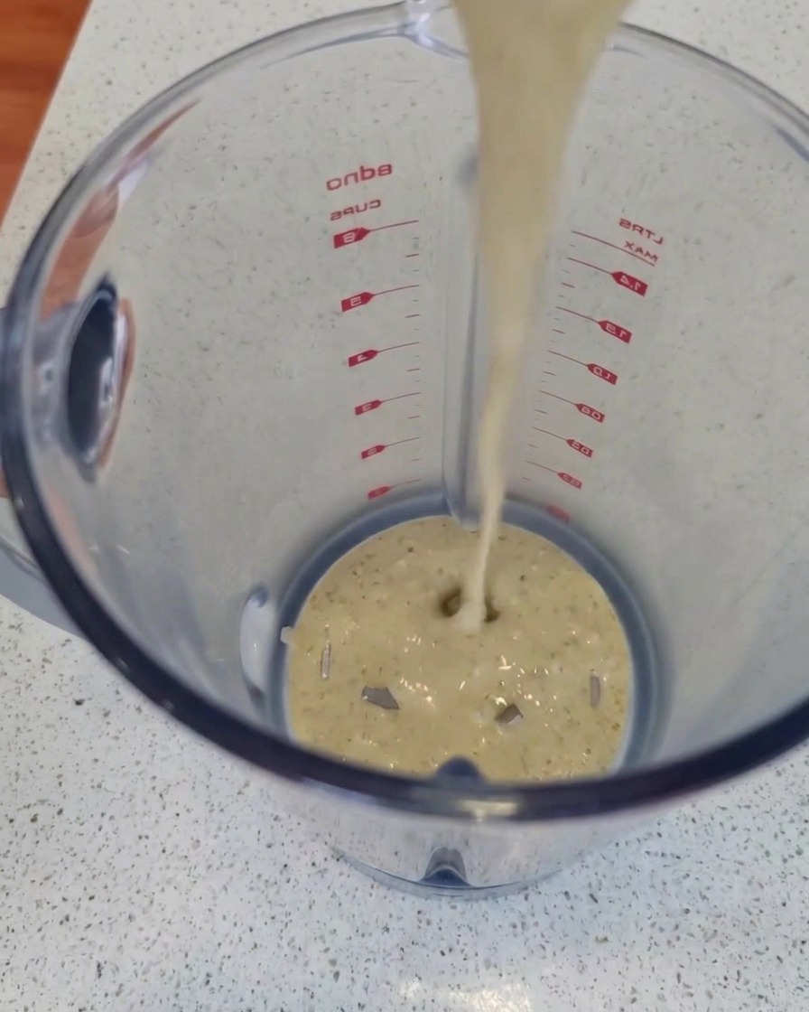 Un filet de sauce crémeuse au bleu parsemée daromates versé den haut dans le bol en plastique transparent dun blender posé sur un plan de travail.