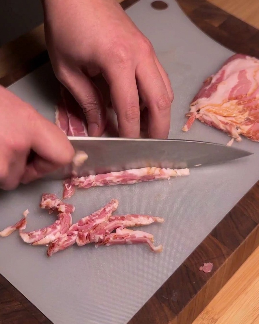 Un chef utilisant un couteau de cuisine bien aiguisé pour couper des tranches de bacon cru en morceaux uniformes sur une planche à découper grise.