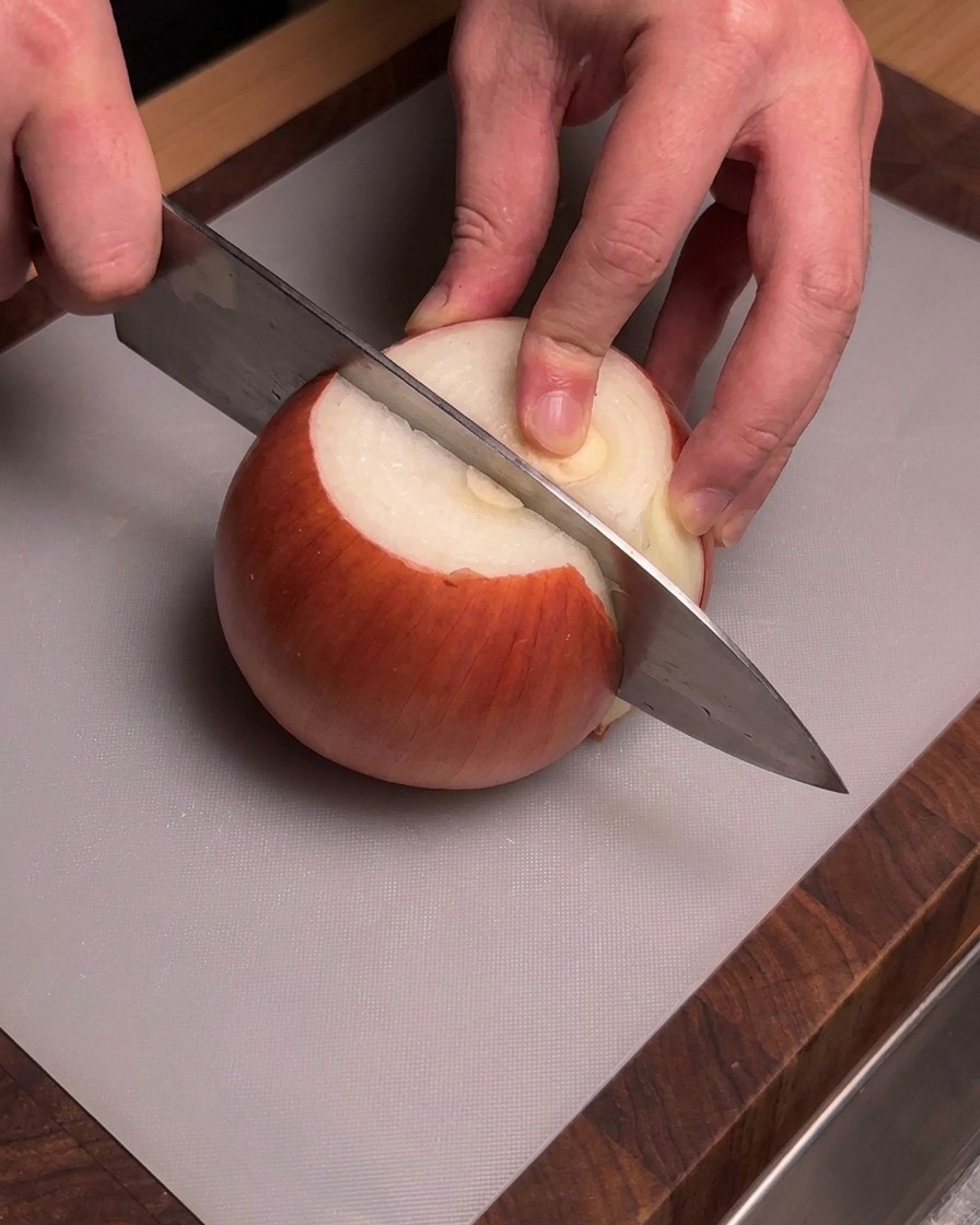 Mains tenant un couteau de chef pour couper un oignon jaune pelé sur une planche à découper grise.