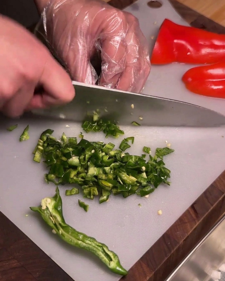 Une personne portant des gants en plastique transparent coupant finement des piments verts ridés épépinés sur une planche à découper.
