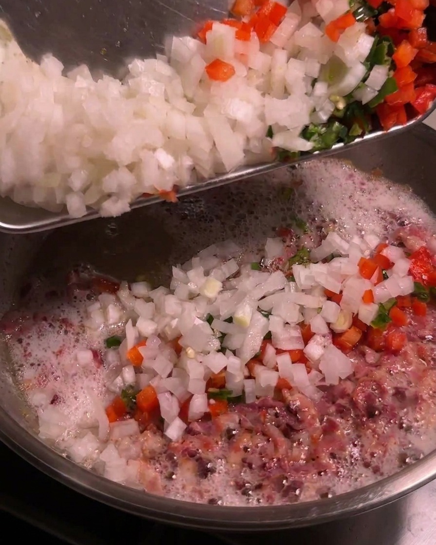 Un plateau contenant des oignons coupés en dés et des piments colorés hachés versés dans une poêle bouillonnante de gras de bacon fondu.