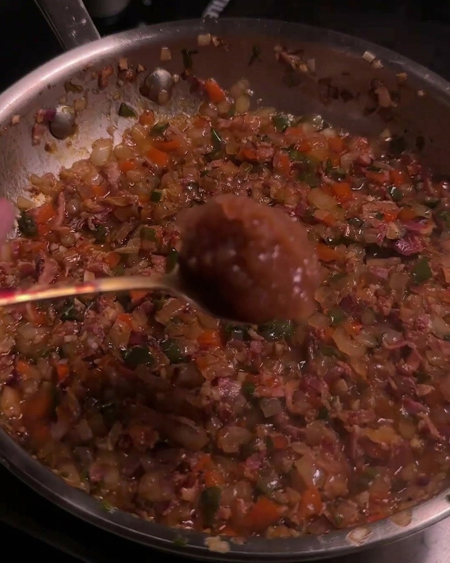 Une cuillère de compote de pommes ajoutée à un mélange caramélisé de bacon et de piments dans une poêle.