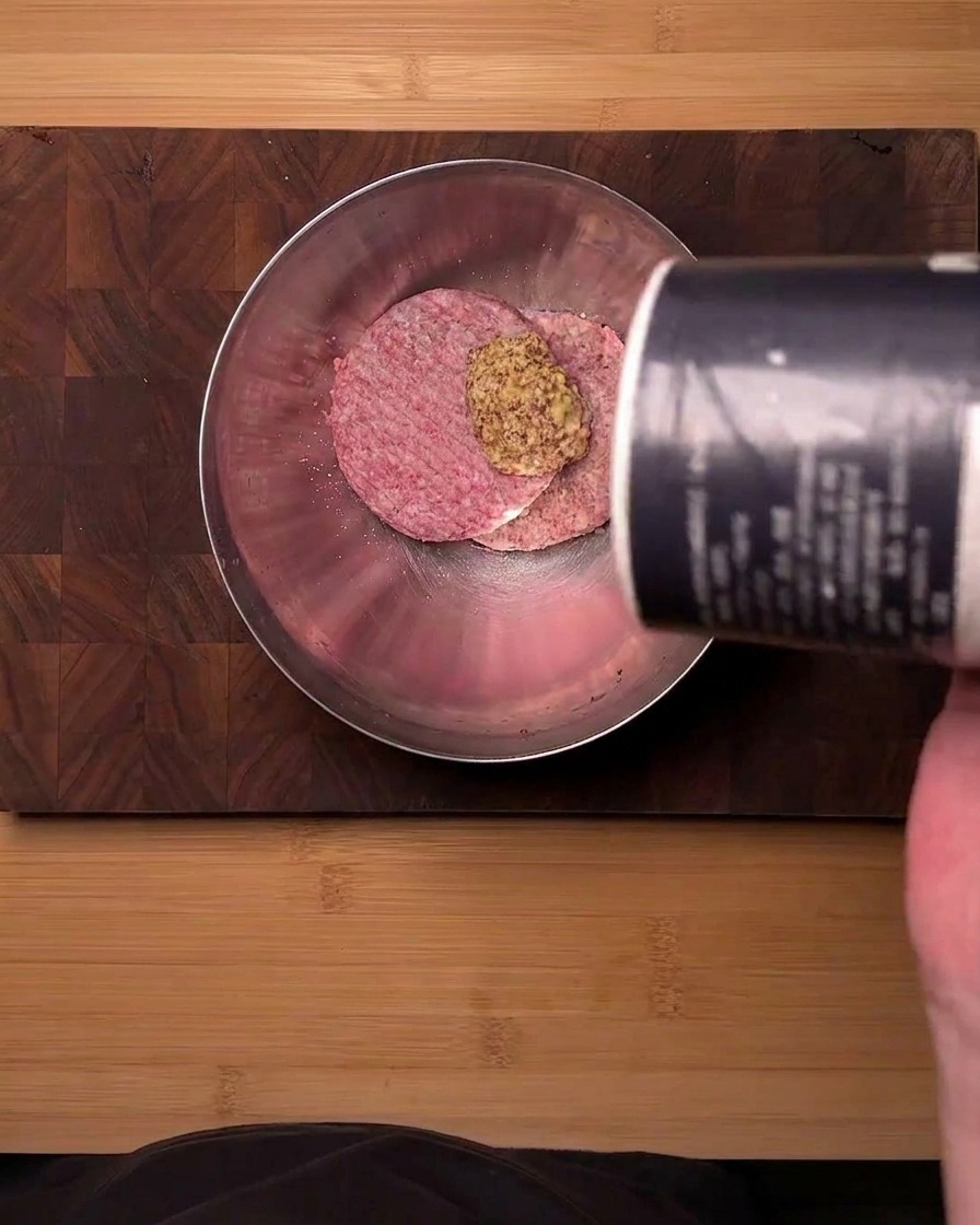 Vue de dessus de bœuf haché cru dans un bol en acier inoxydable assaisonné de moutarde en poudre et de sel versés dun récipient.