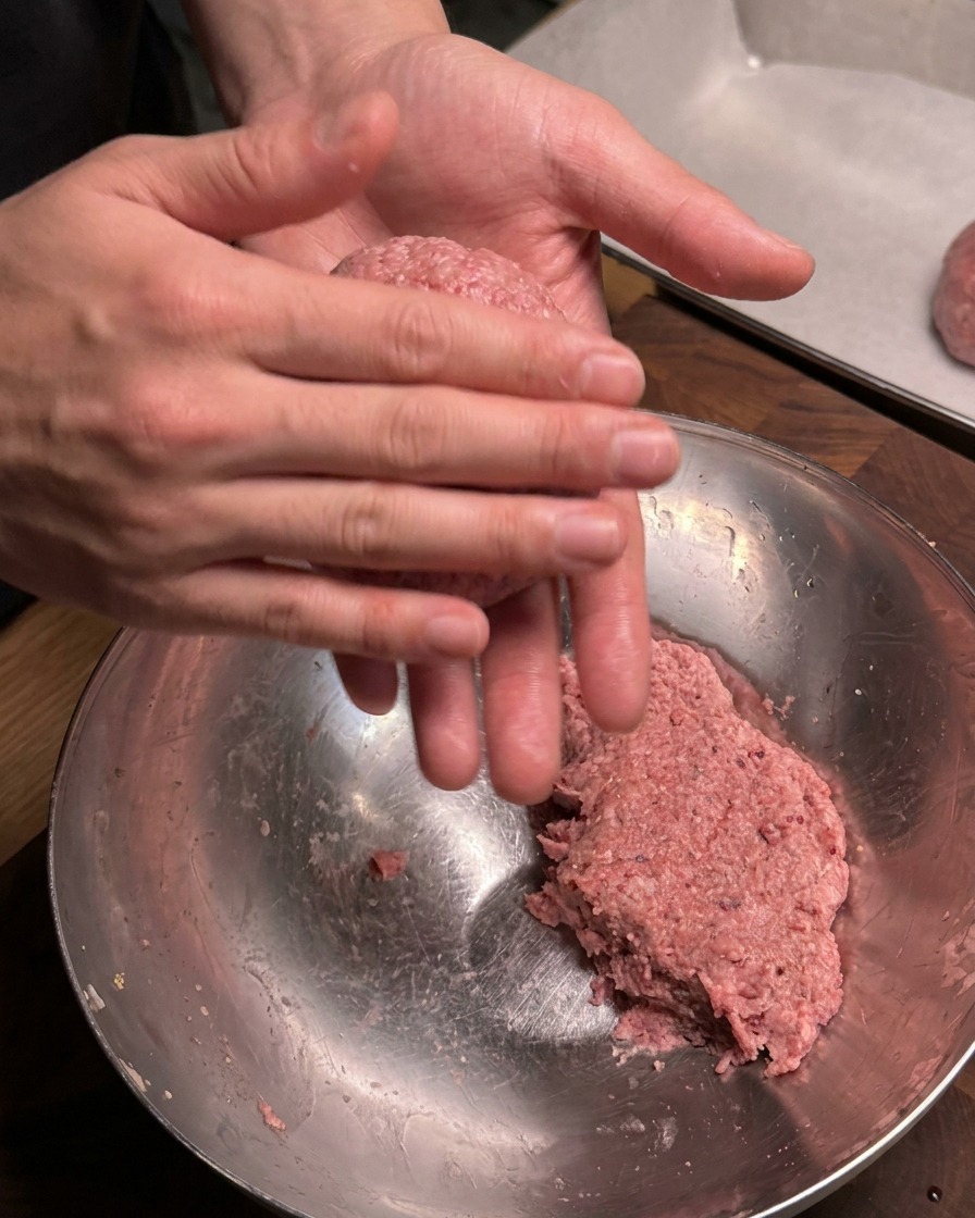 Vue rapprochée de mains roulant et façonnant fermement une portion de bœuf haché cru assaisonné en une boulette ronde au-dessus dun bol de mélange en métal.