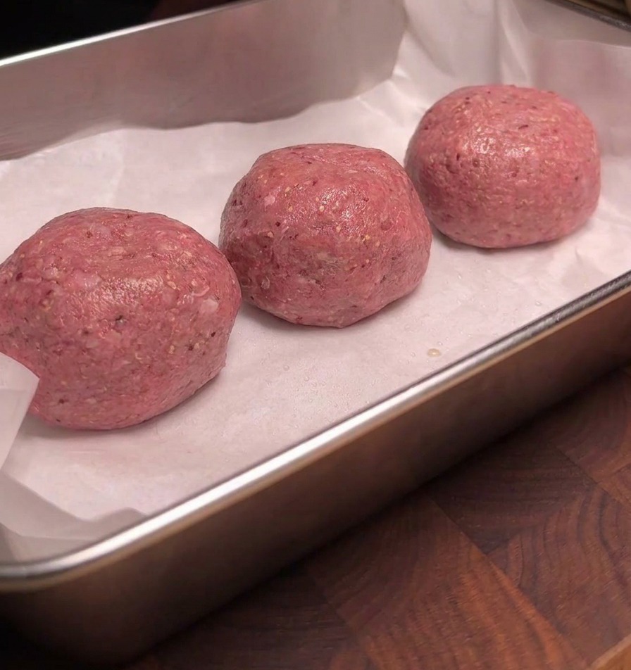 Trois boulettes de bœuf cru disposées proprement sur du papier sulfurisé blanc dans un plateau en métal, leurs surfaces étincelant uniformément dun enrobage de fumée liquide.