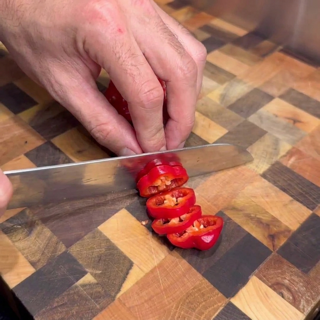 Une personne utilisant un couteau de cuisine pour couper des piments rouges frais en rondelles sur une planche à découper en bois.