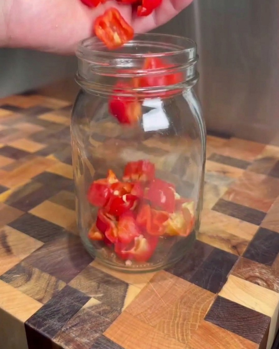 Piments rouges hachés versés dans un bocal en verre transparent posé sur une planche à découper en bois.