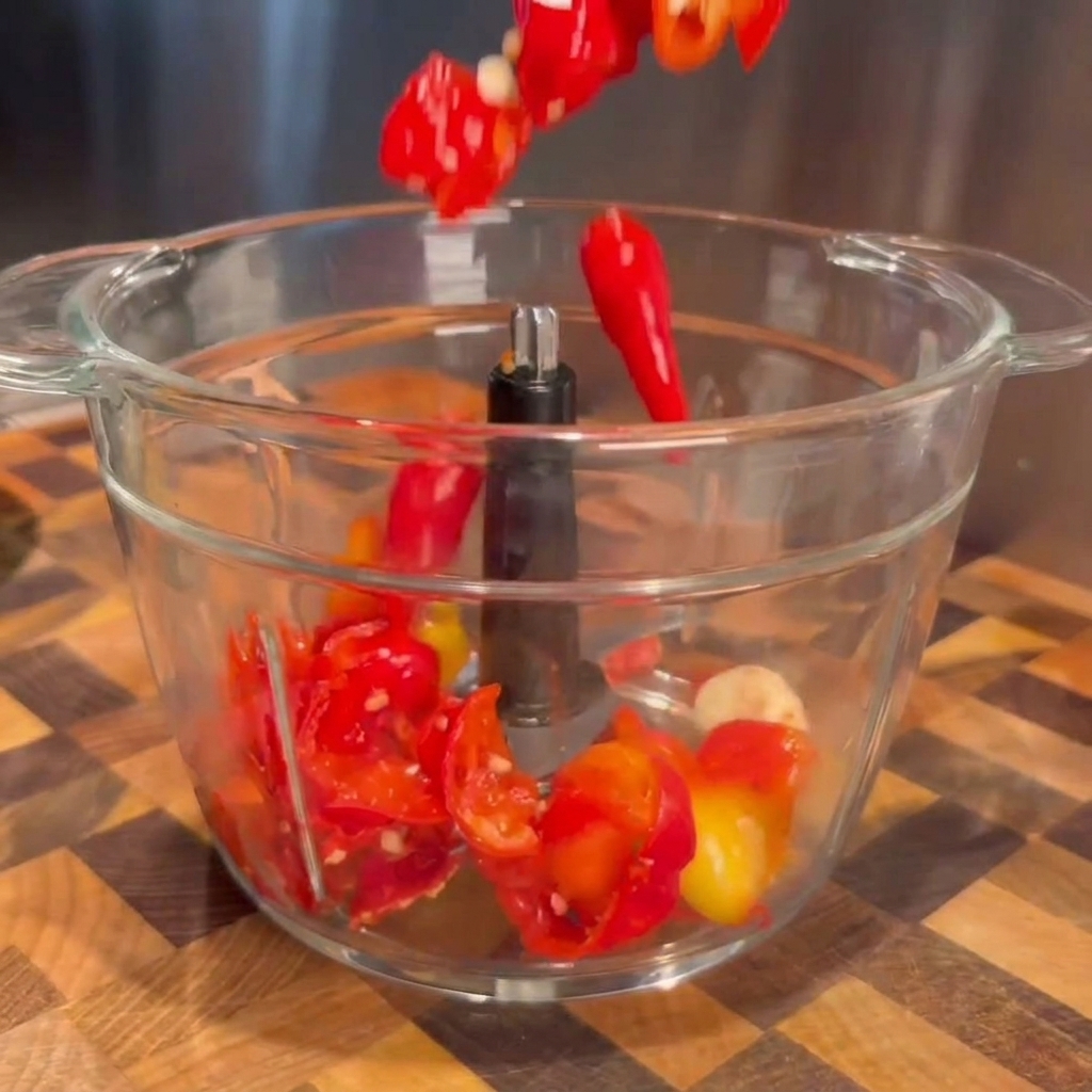 Piments fermentés rouges égouttés et gousses dail tombant dans le bol transparent dun robot culinaire.