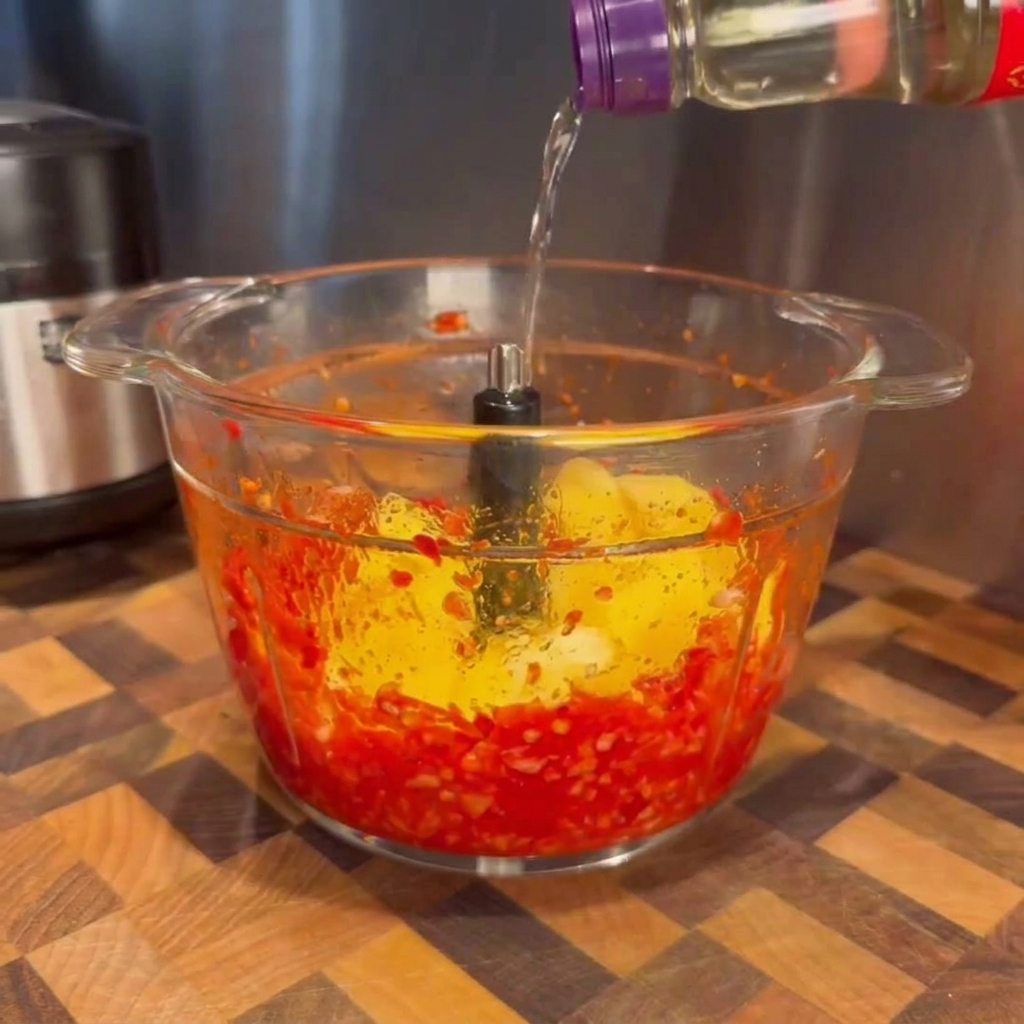 Liquide transparent versé depuis une bouteille dans le bol en verre dun robot contenant des piments rouges mixés et des morceaux de mangue jaune vif.