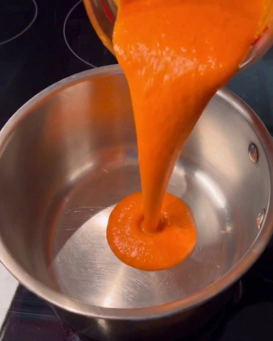 Sauce piquante orange vif coulant de manière fluide depuis un récipient en verre dans une casserole propre en acier inoxydable sur une plaque de cuisson sombre.