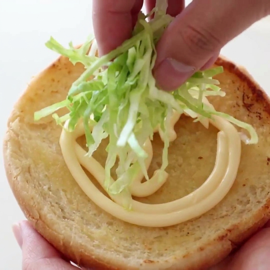 Une main déposant une poignée de laitue croquante sur un pain à burger toasté nappé de mayonnaise.