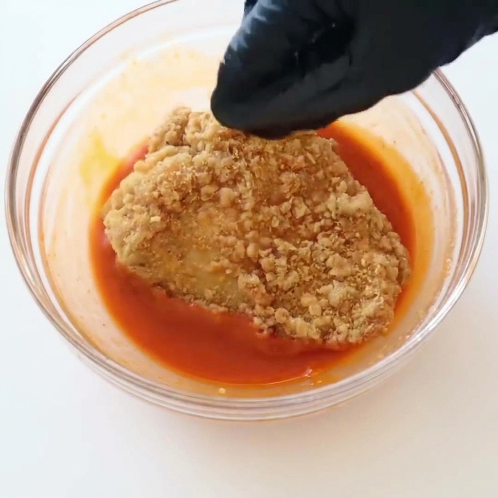Une main gantée de noir plongeant un filet de poulet frit doré dans un bol transparent rempli de sauce buffalo.