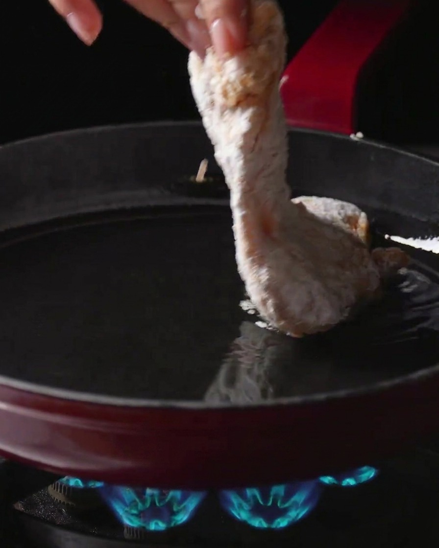 Une main plaçant un haut de cuisse de poulet enrobé de fécule dans une poêle dhuile chaude sur un brûleur à gaz allumé.