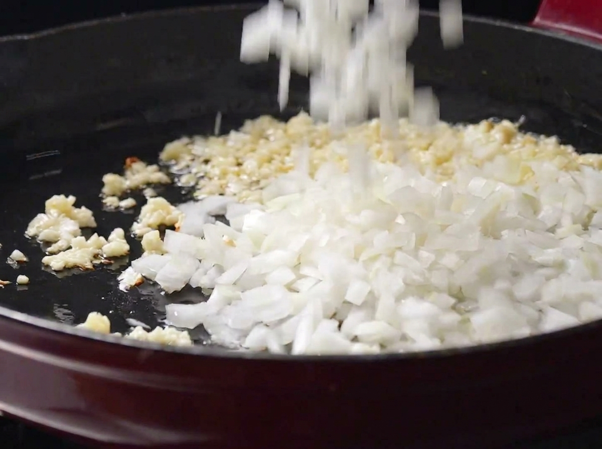 Oignons blancs coupés en dés ajoutés dans une poêle avec de lail haché crépitant.