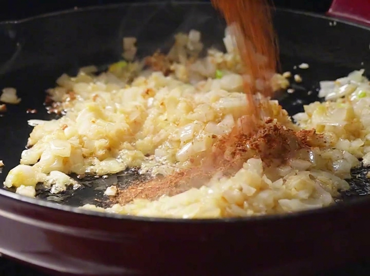 Poudre dépices cajun rouge-brun versée sur des oignons et de lail sautés dans une poêle.