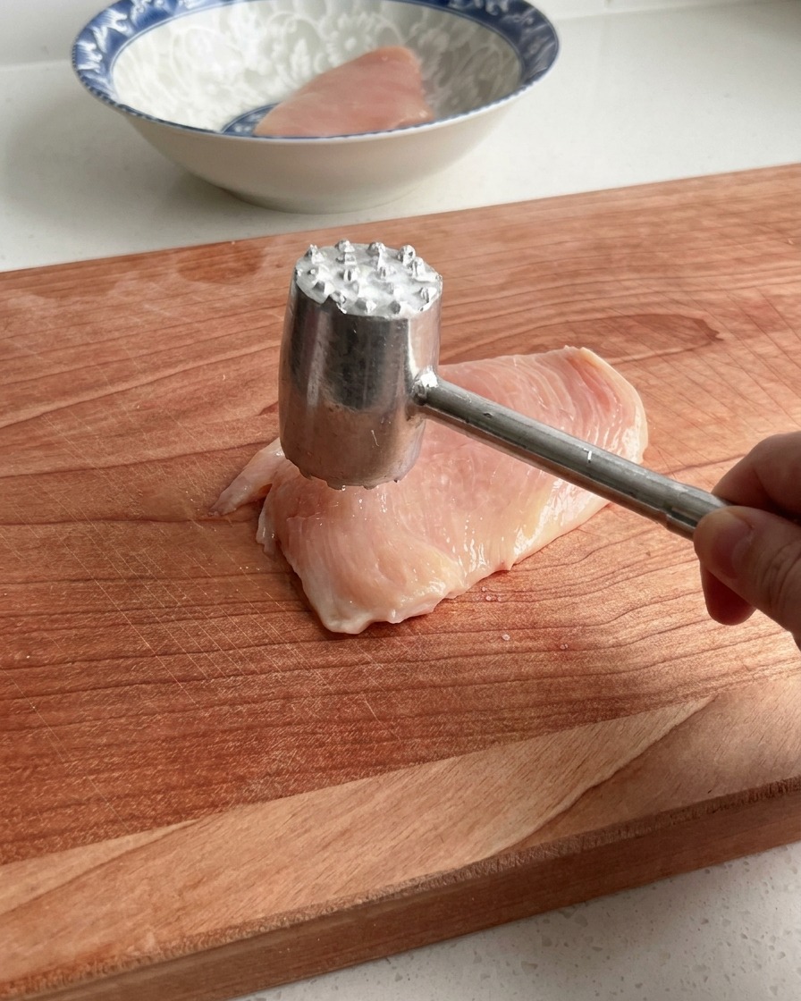Une main utilisant un attendrisseur à viande en métal pour aplatir délicatement un blanc de poulet cru sur une grande planche à découper en bois.