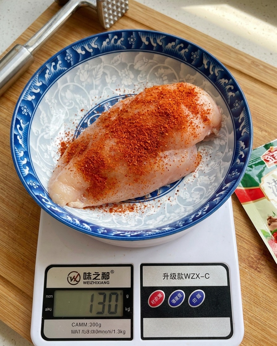 Un morceau de blanc de poulet cru dans un bol en céramique décoré, posé sur une balance de cuisine digitale et généreusement saupoudré dun mélange dépices rouge-orange.