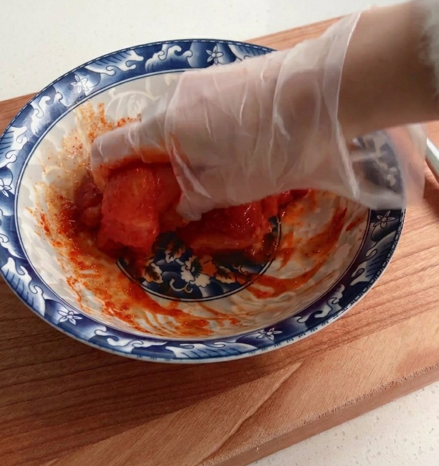 Une main gantée mélangeant soigneusement un blanc de poulet cru avec une épaisse marinade rouge dans un bol à motifs.