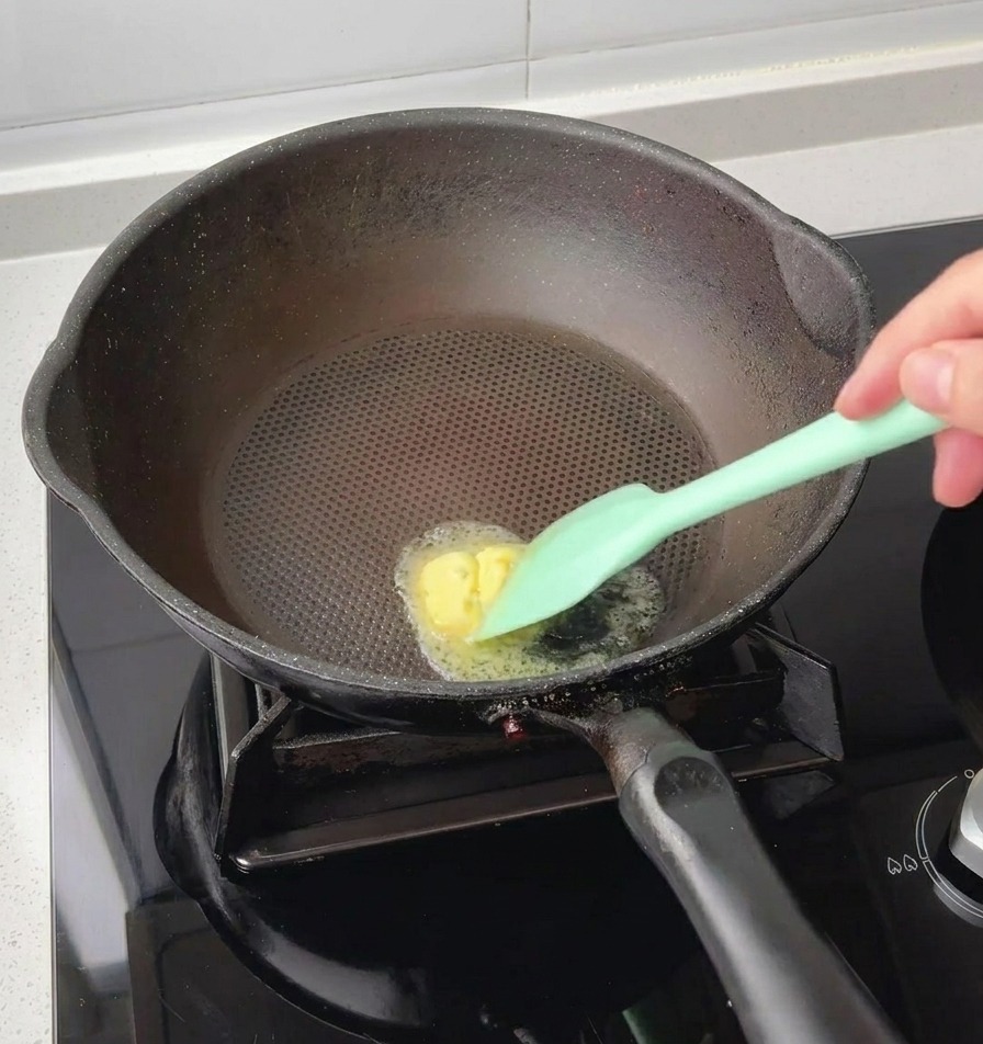 Une spatule en silicone vert faisant fondre une noix de beurre jaune au centre dune poêle noire chaude.