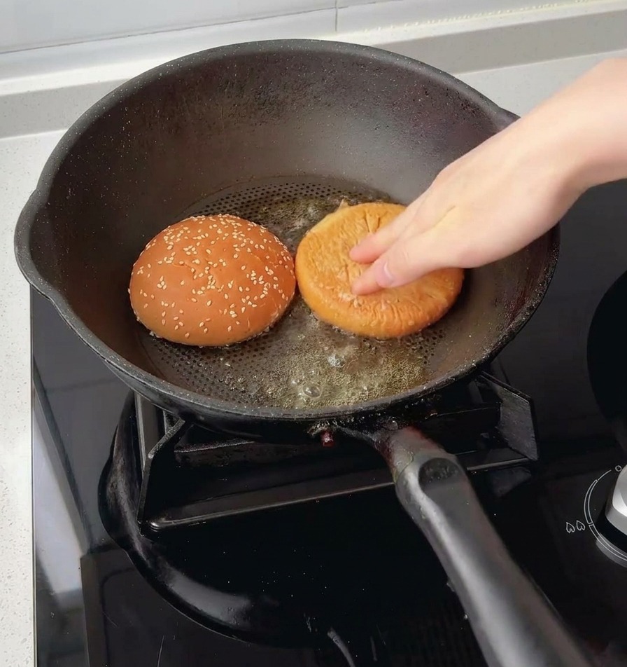 Une main pressant les deux moitiés dun pain à burger au sésame, face mie vers le bas, dans le beurre fondu au fond dune poêle.