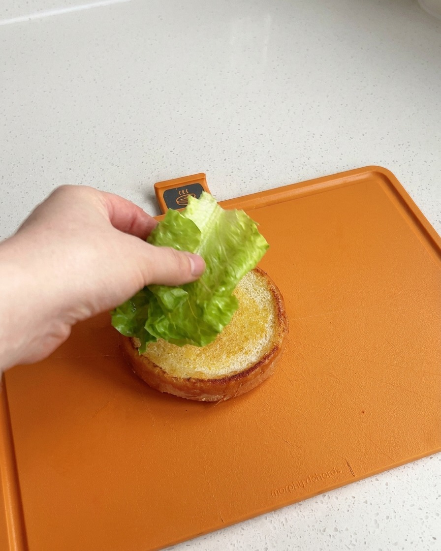 Une main déposant une feuille de laitue verte et bien propre sur la base dorée et toastée dun pain à burger, posé sur une planche à découper orange.